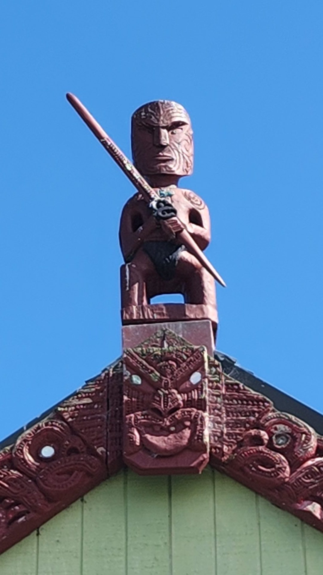 Carved figure on the wharenui roof against a blue sky.